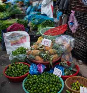 A vibrant market stall displays a variety of fresh produce. There are baskets filled with limes, pineapples, cucumbers, and other leafy greens. Plastic bags, crates, and sacks are scattered around, indicating an active trading area. Price tags are visible on some products.